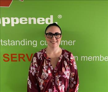 Female employee Sarah Raymond standing in front of a green wall with the SERVPRO mission statement